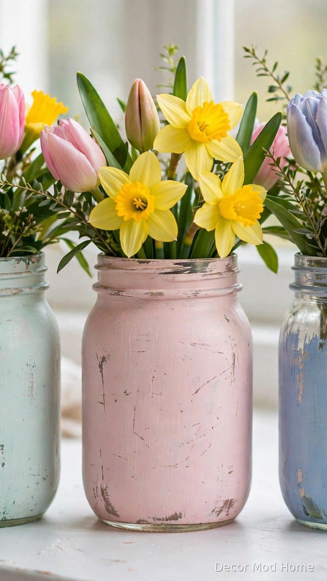 Mason jars hand-painted in pastel mint, pink and blue holding tulips and daffodils on a windowsill